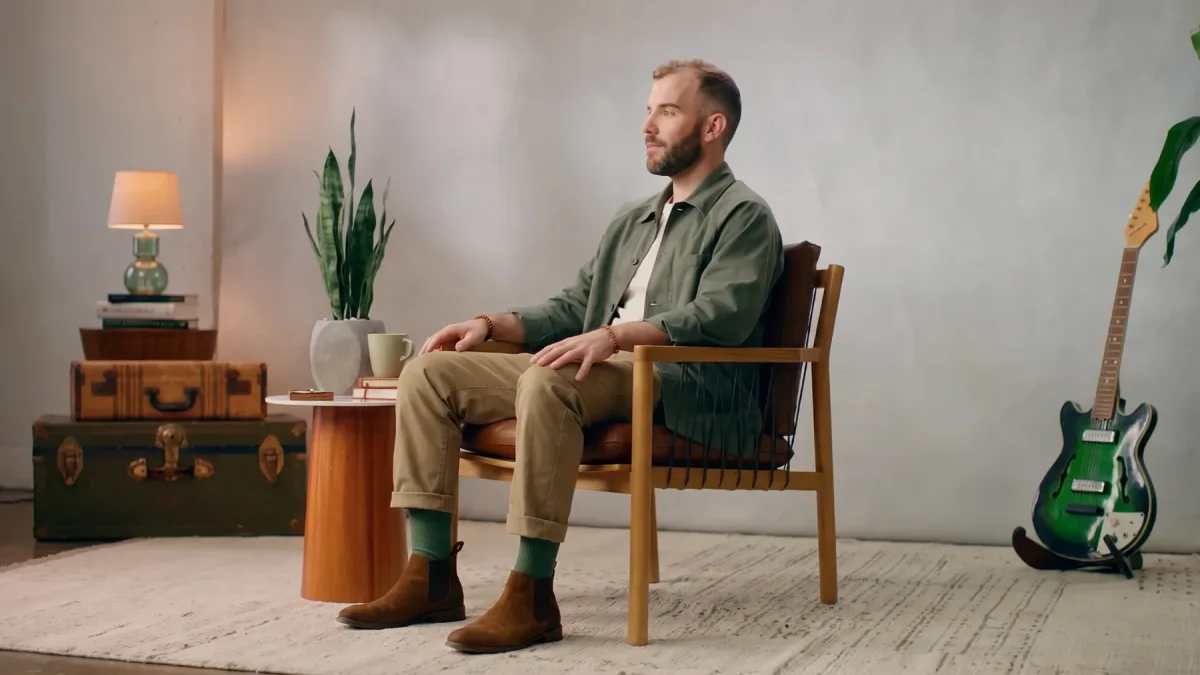 Matthew Karsten seated in a wooden chair in the studio, with a guitar and a side table, wide shot.